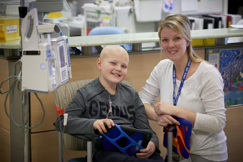 Chase sits on a chair holding a tablet with a chemotherapy pump to his right. To his left is child life specialist, Courtney Knight. Both smile and pose for a photo.