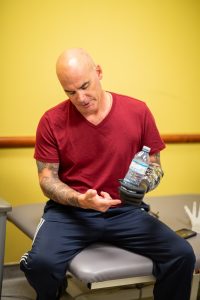 Craig sits on a bench in a clinical room holding a water bottle with his myoelectric hand