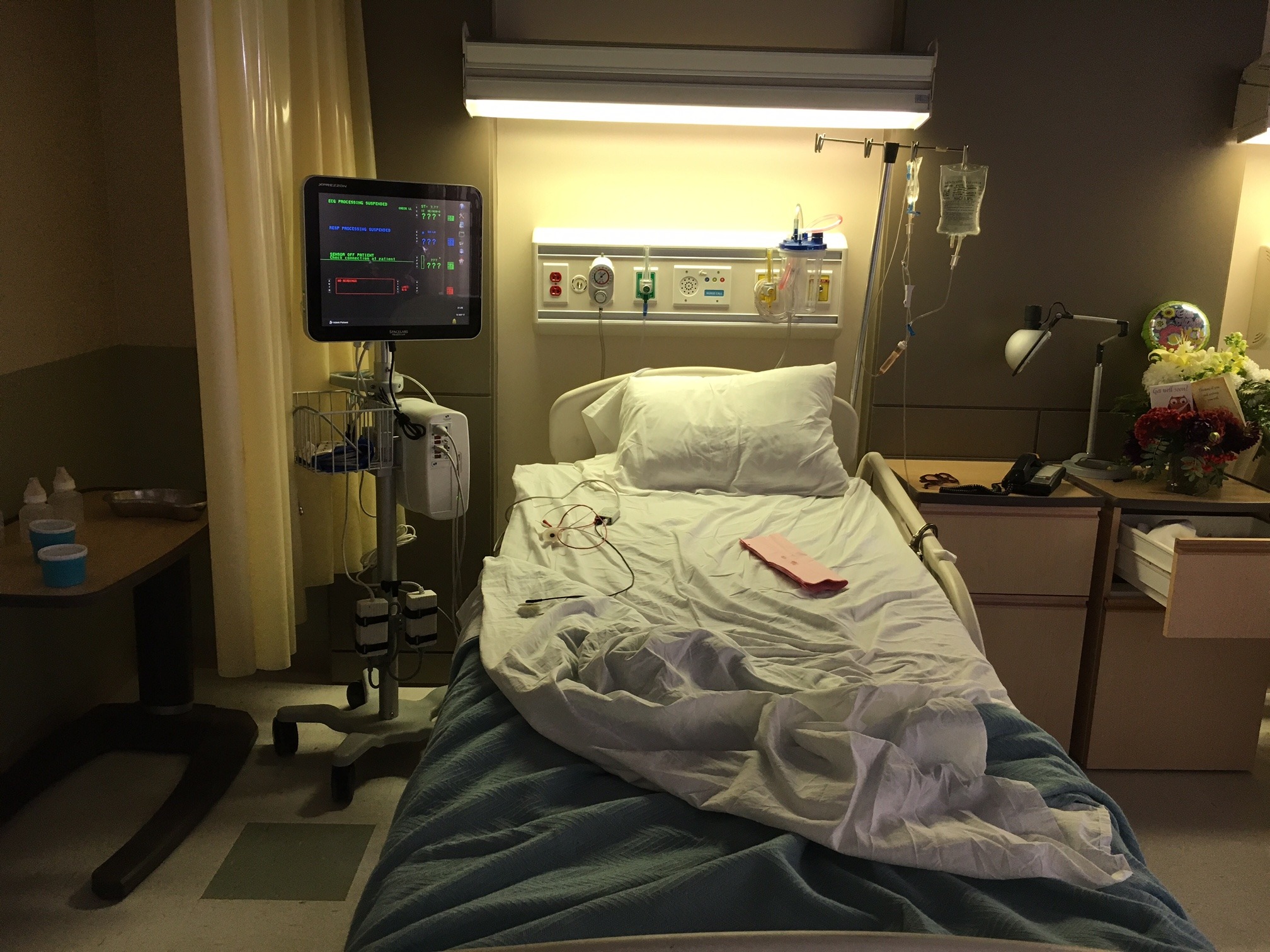 A hospital room including a bed, light, monitor and table with balloons and flowers