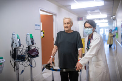 Dr. Mylinh Duong walks with an elderly patient using a walker along a hospital hallway.