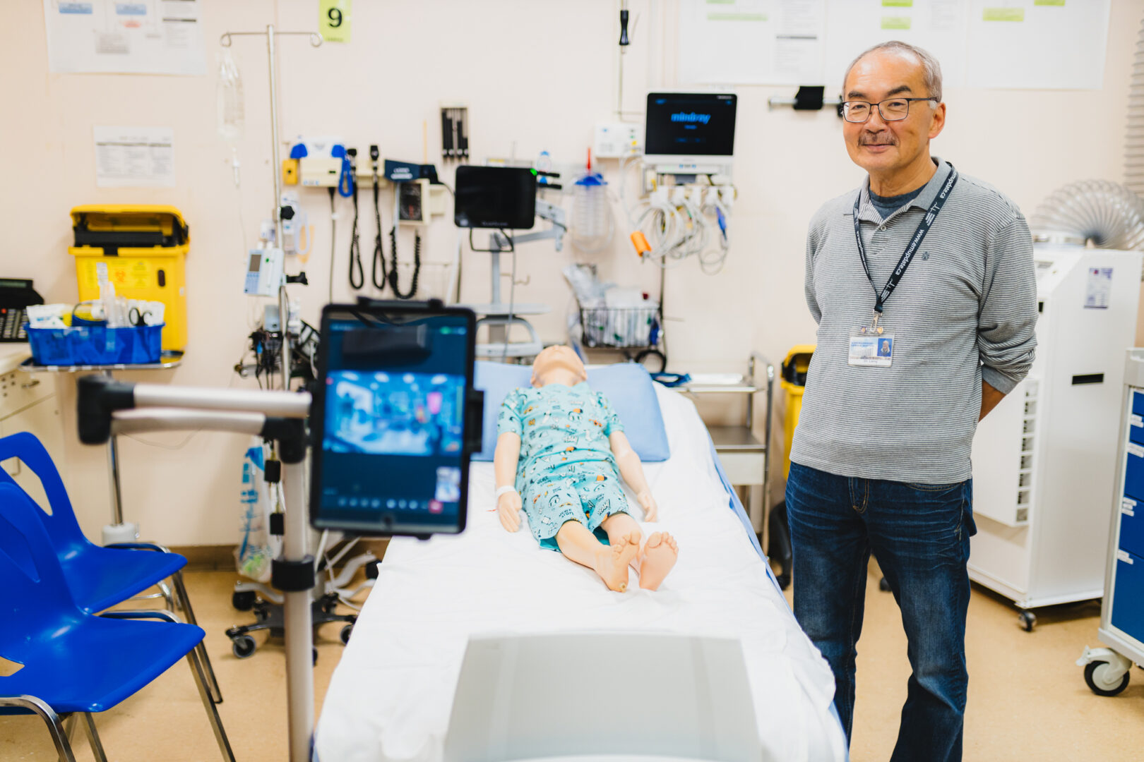 Dr. William Liang stands beside a mannequin on a hospital bed. In the foreground is an iPad on a stand, set up with video chat with the mock patient and doctor on the screen.