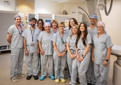 A group photo of 11 health care workers wearing scrubs while standing in a hospital procedure room.