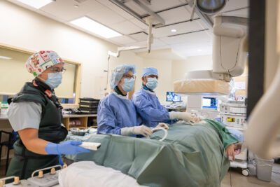 Three health care providers in caps, masks, gowns and gloves look up at a screen. A mock patient lies on a table in front of them, under a sheet.