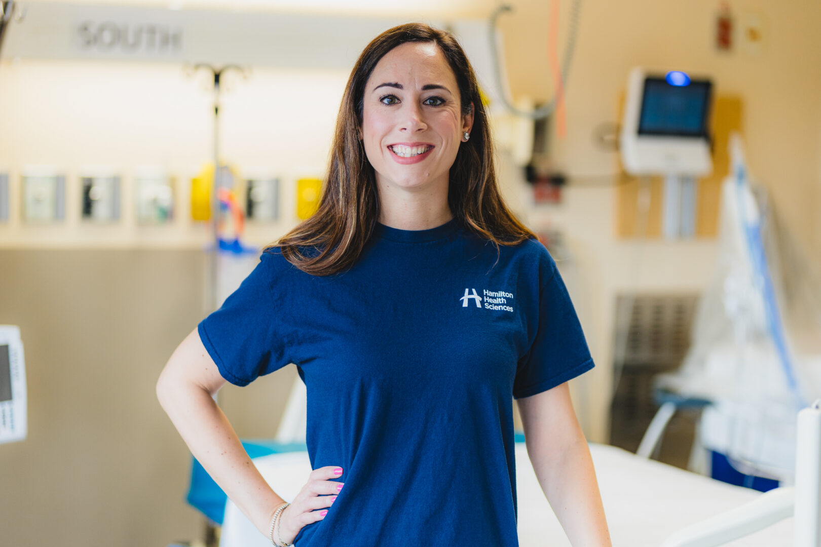 Patient researcher Barbara Dolanjski stands in a hospital room, smiling, wearing a dark blue HHS T-Shirt