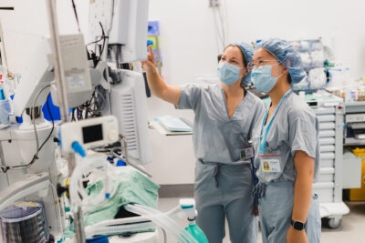 Grade 12 graduate Stephanie Chan and HHS research coordinator Jessy Moore are in a hospital operating room, dressed is hospital scrubs, caps and masks